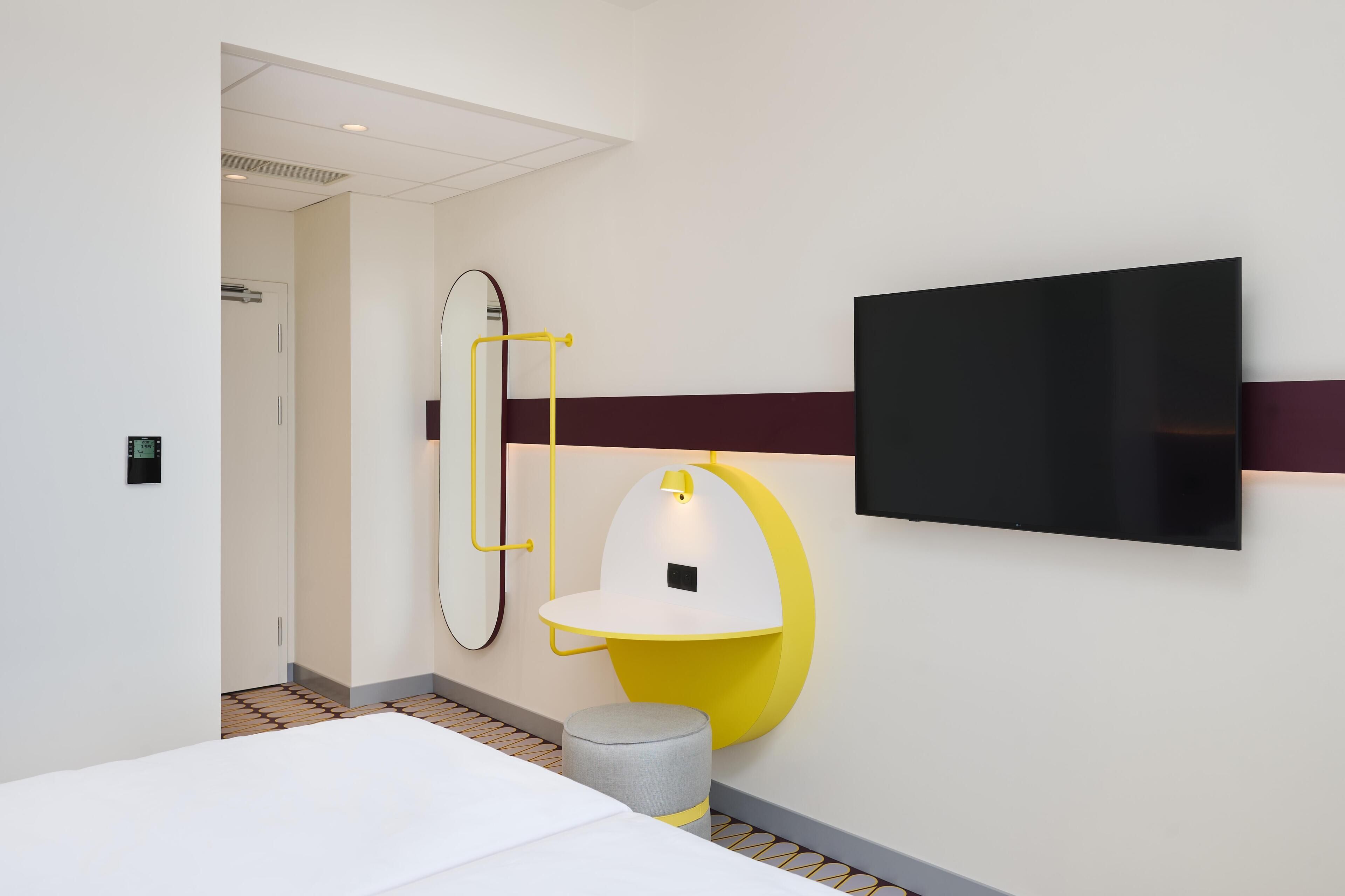 A modern hotel room with a bed, a wall-mounted TV, a mirror, and a unique yellow and white circular light fixture.