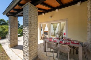 An outdoor patio with a stone column, a table set for dining, wicker chairs, and a view into a room with a sliding glass door. - Villa Vinea (100691-K1) - Momjan (Momjan)