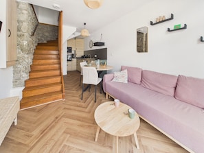 A modern interior with a herringbone wooden floor, a pink sofa, a round wooden table, and a kitchen in the background. - Stay for 2, walk everywhere, beach 500m (Dinard)