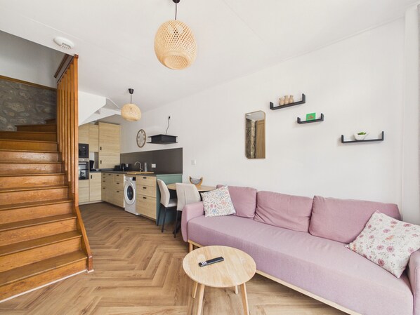 A modern living room with a wooden staircase, a sofa, a round wooden coffee table, and a kitchen in the background. - Stay for 2, walk everywhere, beach 500m (Dinard)