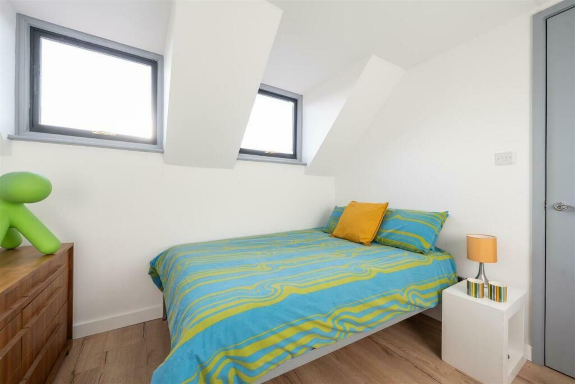 Une chambre à coucher avec un lit, deux fenêtres, une commode, une lampe et une porte.