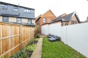A residential backyard with a wooden fence, a grassy area, and a white wall. - Contemporary 3BDR House in South Woodford (Greater London)