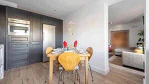 A modern dining area with a wooden table set for four, a kitchen with dark cabinetry, and a living space with a sofa and a plant. - Contemporary 3BDR House in South Woodford (Greater London)