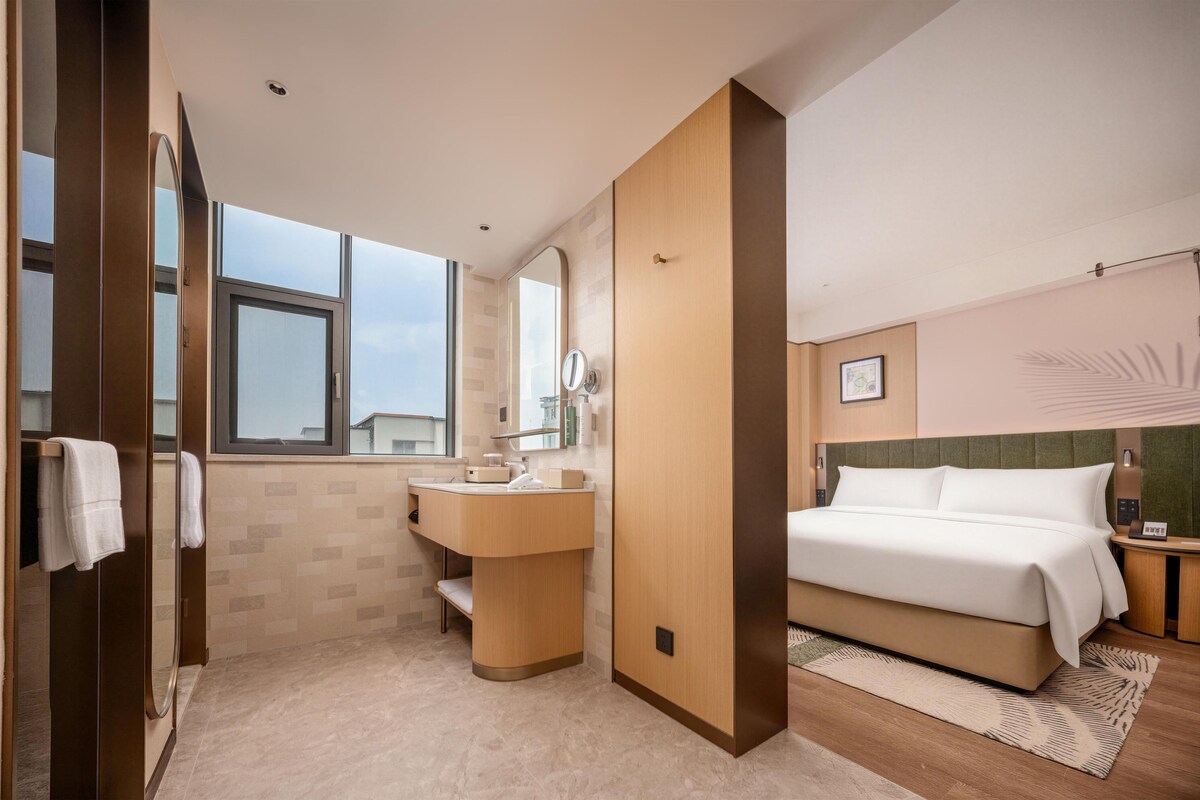 Quarto de hotel moderno com uma cama grande, banheiro com toucador e janela com vista.