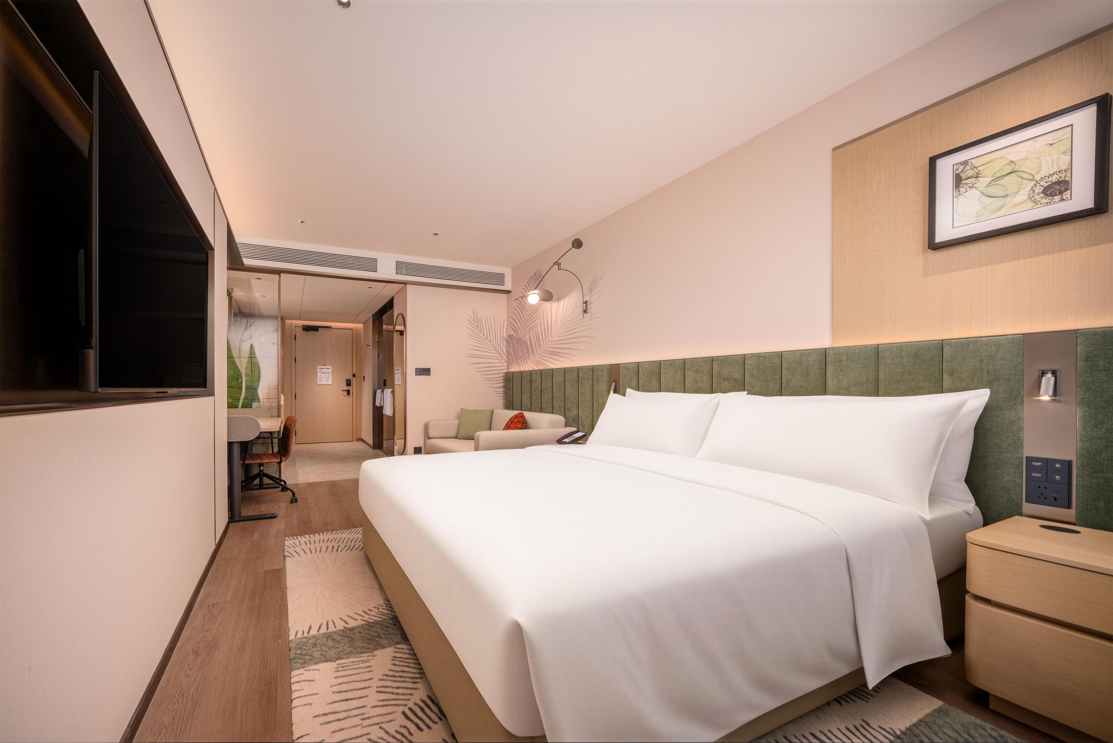 Une chambre d’hôtel comprenant un lit, une table de chevet, un bureau avec une chaise, un téléviseur fixé au mur et un tableau encadré.