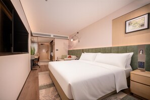 Quarto de hotel com cama, mesa de cabeceira, escrivaninha, cadeira, televisão presa à parede e um quadro emoldurado.