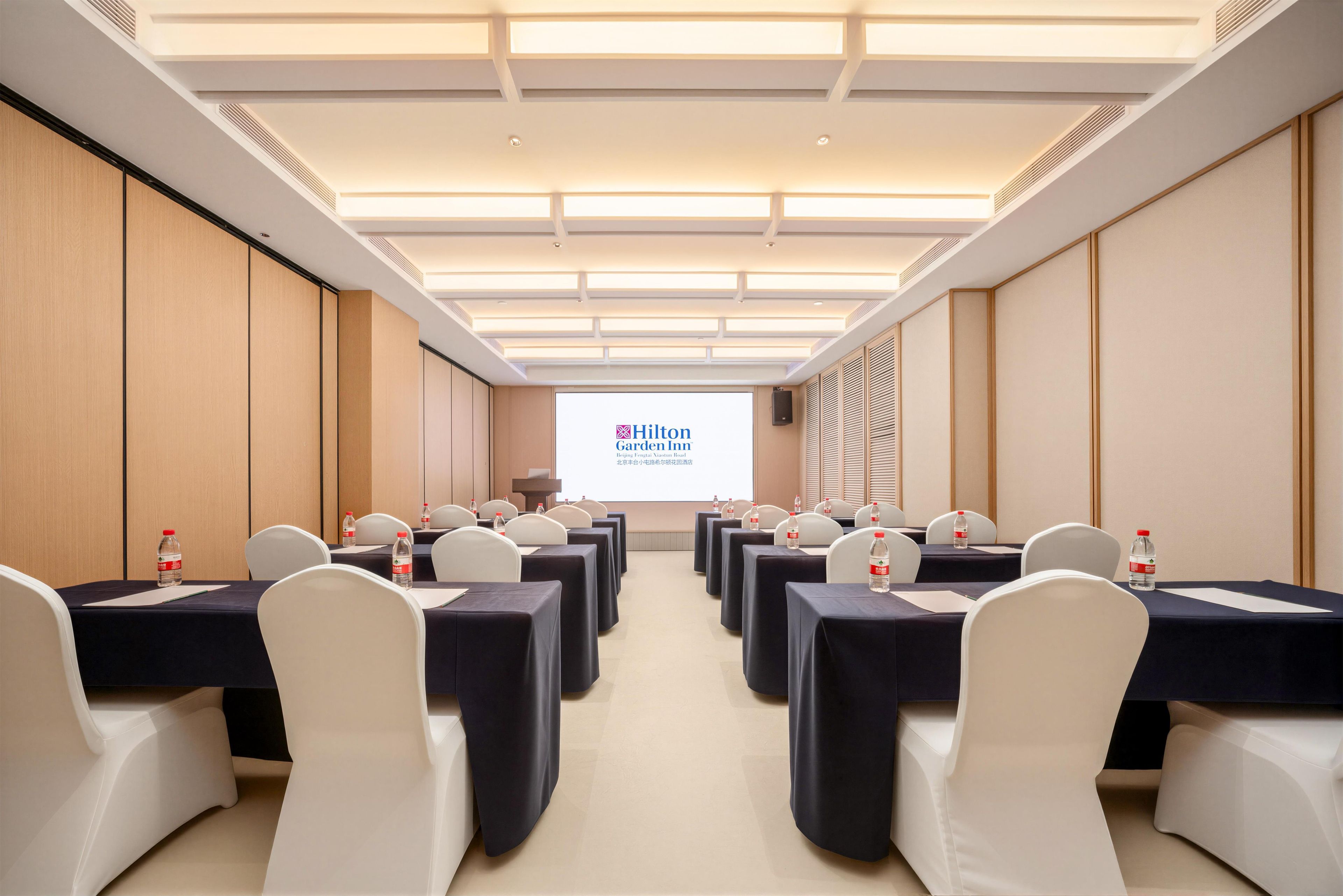 Un salón de conferencias con filas de mesas y sillas, una pantalla de proyector mostrando el logotipo de Hilton Garden Inn, y un altavoz en la pared.