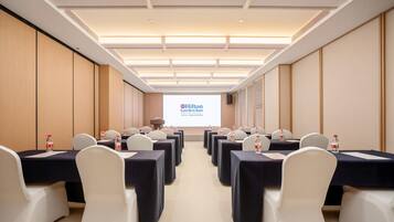 Un salón de conferencias con filas de mesas y sillas, una pantalla de proyector mostrando el logotipo de Hilton Garden Inn, y un altavoz en la pared.