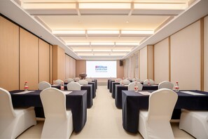Uma sala de conferências com fileiras de mesas e cadeiras, uma tela de projetor exibindo o logotipo do Hilton Garden Inn e um alto-falante na parede.