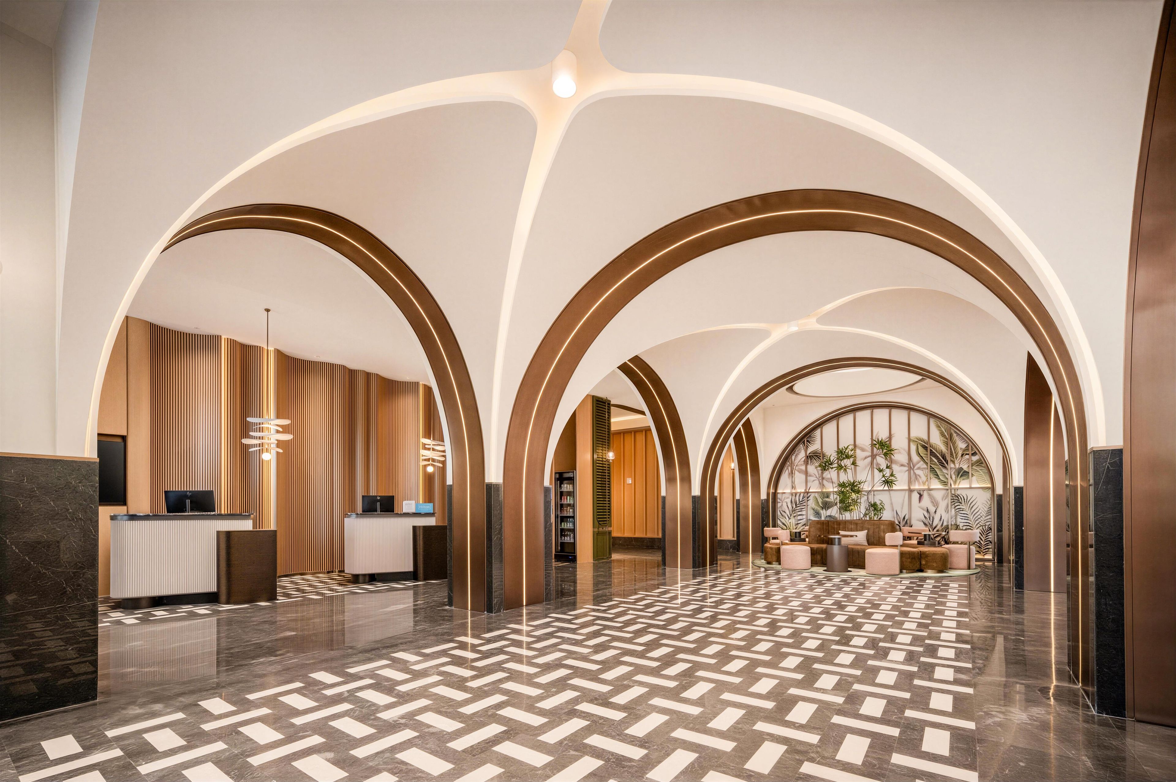 A spacious lobby with a geometric patterned floor, a reception desk, and a seating area with a view of indoor plants.