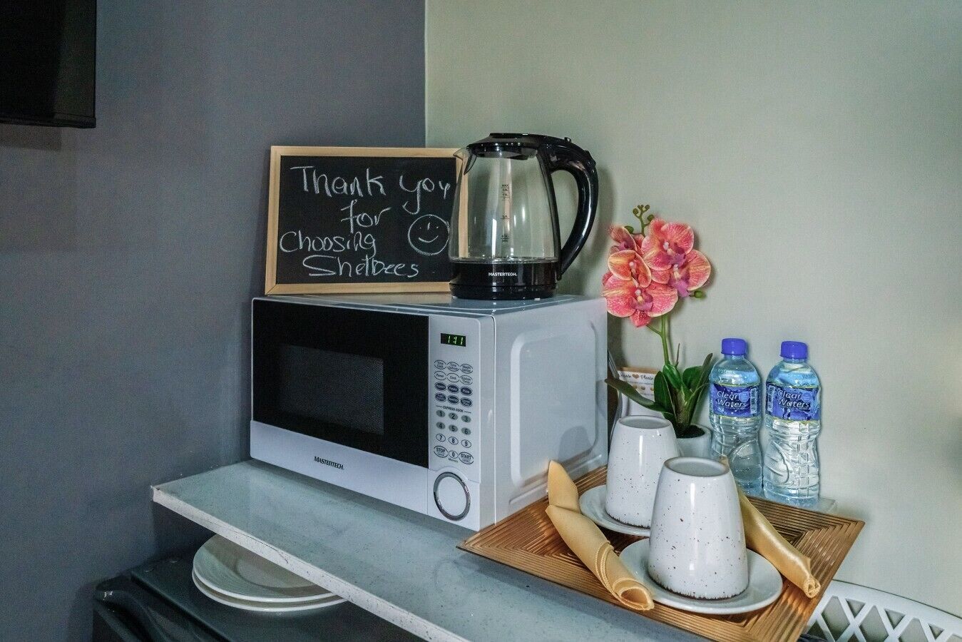 Un coin cuisine équipé d’un four à micro-ondes, d’une machine à café, de bouteilles d’eau et d’un tableau noir sur lequel est écrit un message.