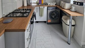 A compact kitchen with a gas stove, a built-in washing machine, and a window providing natural light. - Home -near Town/lutonairport Sleeps 6/parking/wifi (Luton)