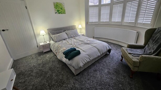 A bedroom with a bed, two bedside lamps, a radiator, a window with blinds, and a chair. - Home -near Town/lutonairport Sleeps 6/parking/wifi (Luton)
