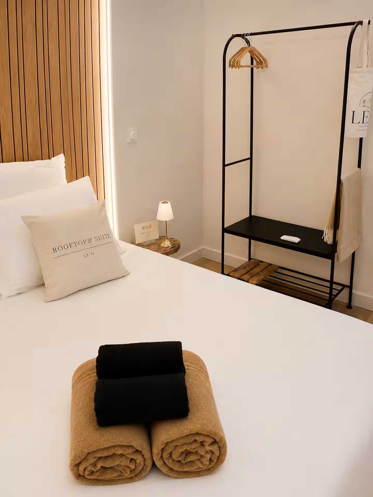 Quarto de hotel com uma cama, uma mesa de cabeceira com abajur, um guarda-roupa de metal preto e uma toalha sobre a cama.