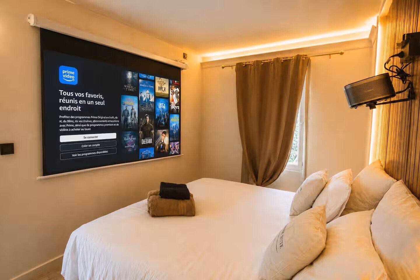 Une chambre d’hôtel dotée d’un grand écran affichant l’interface de Prime Video, d’un lit avec une literie blanche, d’un petit tabouret en bois et d’une fenêtre avec des rideaux.