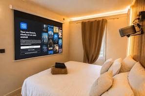 Habitación de hotel con una pantalla grande mostrando la interfaz de Prime Video, una cama con ropa blanca, un taburete de madera pequeño y una ventana con cortinas.