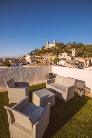 A rooftop terrace with wicker furniture, a clear view of a historic castle, and a town below.