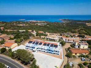 Vue aérienne d’un quartier résidentiel situé sur la côte, avec des maisons, un grand bâtiment doté de panneaux solaires et une vue dégagée sur la mer.