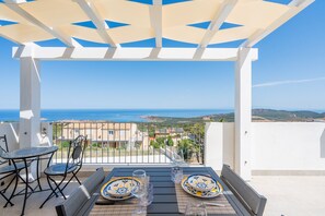 Ein Balkon mit einem für zwei Personen gedecktem Tisch, der einen malerischen Blick auf das Meer und entfernte Gebäude bietet.