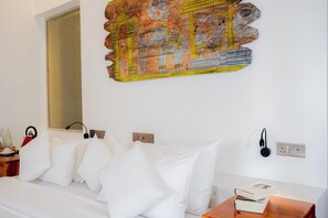 A hotel room with a bed, a wall-mounted artwork depicting a historical scene, a bedside table with a kettle, and a bedside lamp. - Thaulle Luxury Boutique & Social Club  (Kirinda)