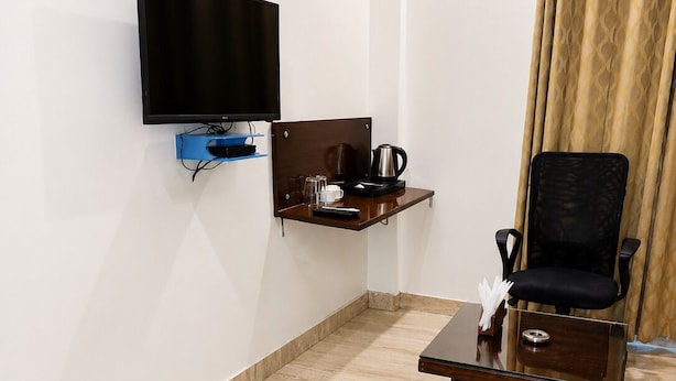 A hotel room with a flat-screen TV mounted on the wall, a small wooden side table with a coffee pot, a chair, and a glass cabinet. - HOTEL K11 (Haldwani)