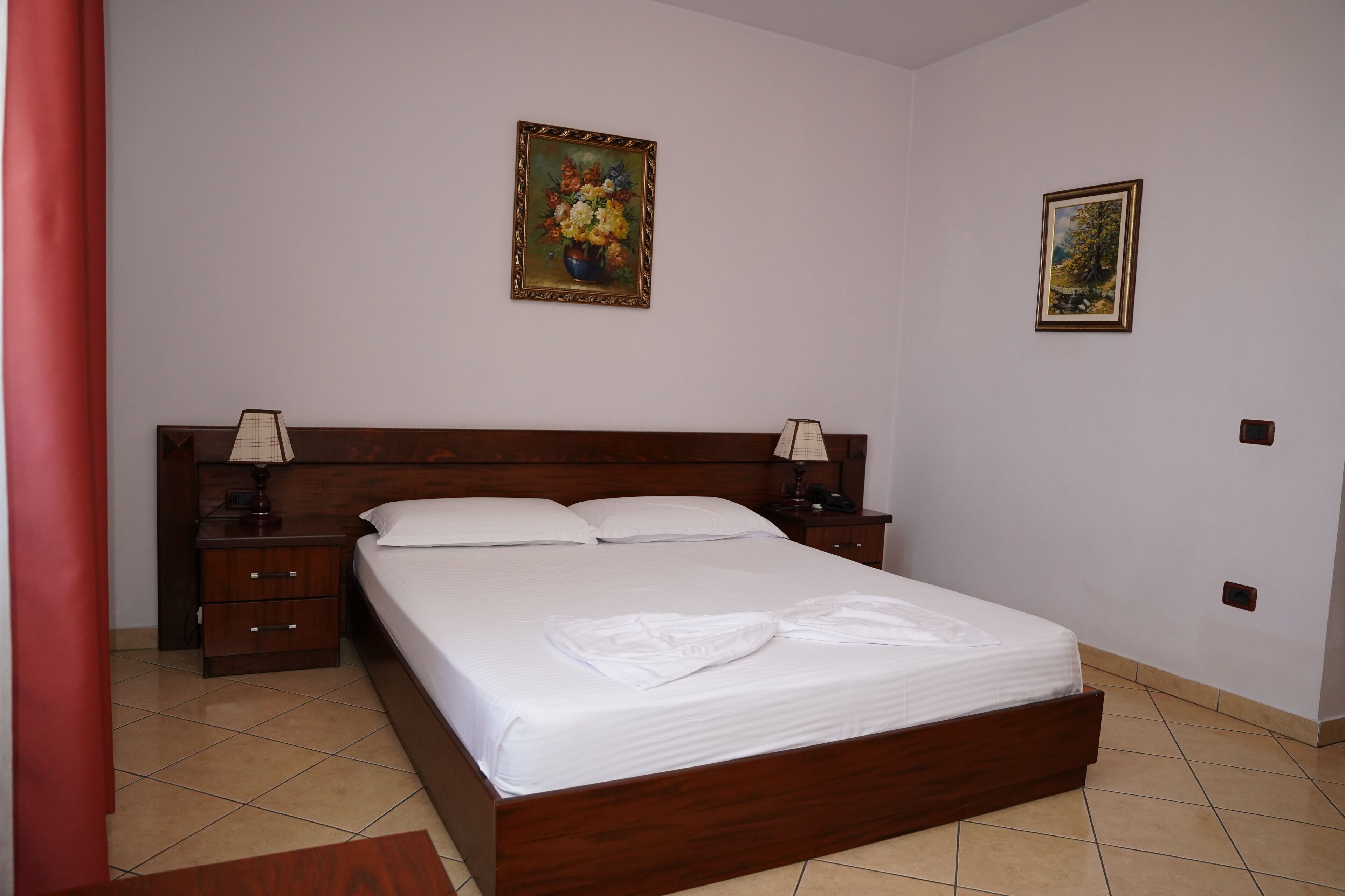 Una cama bien tendida en una habitación de hotel con cabecera de madera, dos mesitas de noche con lámparas y dos cuadros enmarcados en la pared.