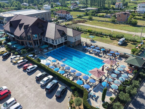 A swimming pool with multiple lanes, surrounded by lounge chairs and umbrellas, and a parking area with several cars.