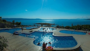 Un espace piscine comprenant plusieurs piscines, entouré d’une terrasse aménagée et offrant une vue sur une étendue d’eau et des reliefs lointains.