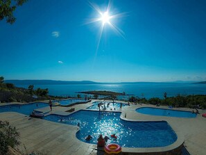 Ein Poolbereich mit mehreren Schwimmbecken, umgeben von einer angelegten Terrasse, mit Blick auf ein Gewässer und entfernte Landschaften.