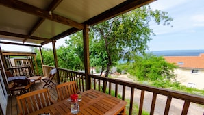 Une terrasse en bois avec une table et des chaises, offrant une vue panoramique sur un paysage verdoyant, avec des arbres et la mer à l’horizon.