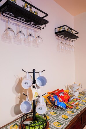 Una habitación con una estantería de pared para vinos con varias copas, un soporte con tazas y un hervidor, y una mesa con aperitivos y un mantel estampado.