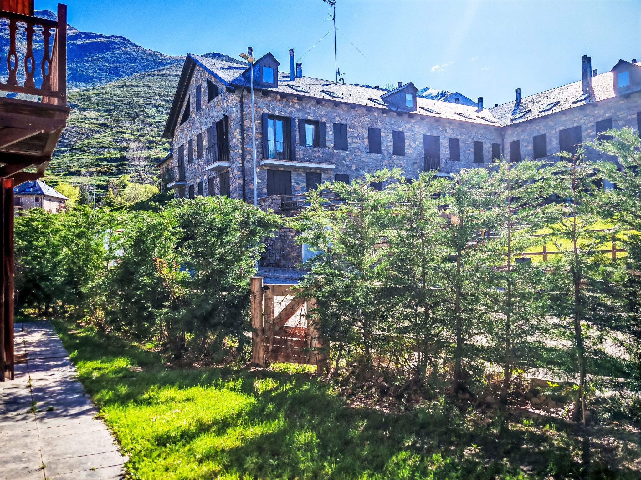 A stone building with multiple windows and a balcony, surrounded by greenery and a mountain in the background.