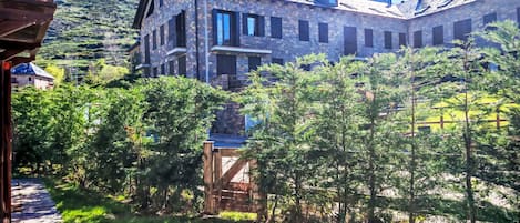 A stone building with multiple windows and a balcony, surrounded by greenery and a mountain in the background.