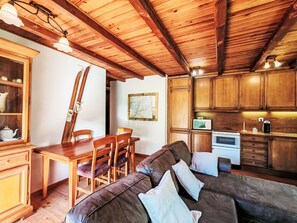 Een gezellige zithoek met houten plafond, een eettafel met stoelen, een bank met kussens, en een keuken met kasten en apparatuur.