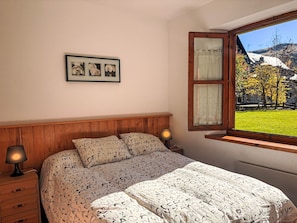 A bedroom with a bed, a nightstand, a lamp, a window with a view of a garden, and a framed picture on the wall. - Bajo con jardín y parking - Dellui 2 by Interhome (Taüll)