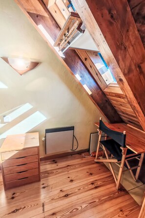 Um quarto com teto inclinado, vigas de madeira, claraboia, radiador, cómoda de madeira e uma cadeira dobrável.