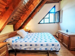 Um quarto com teto inclinado, uma cama com colcha estampada, uma mesa de cabeceira com abajur, um armário de madeira e uma janela com vista.