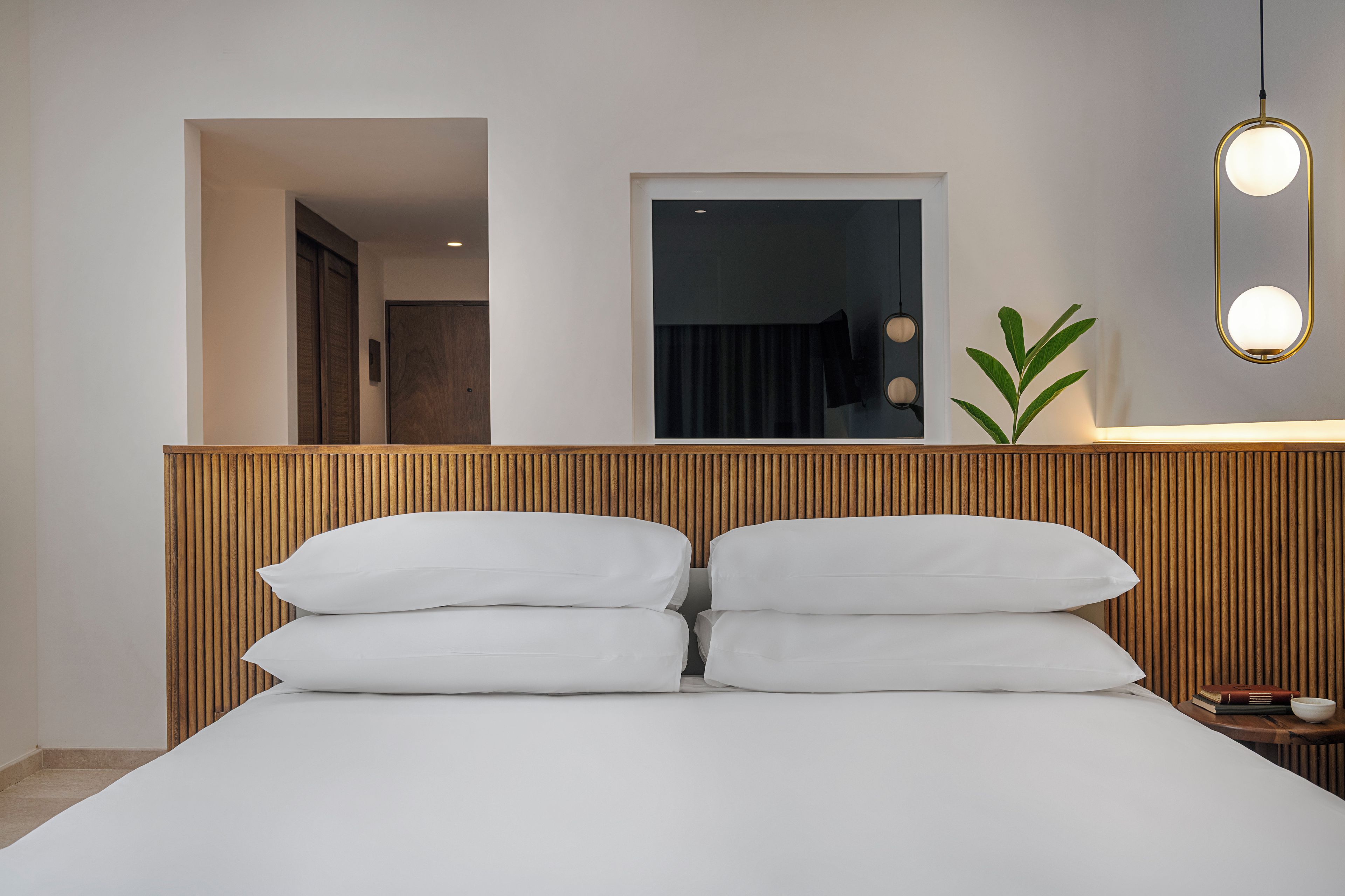 A hotel room with a wooden headboard, a bed with white linens, a bedside table with a book and a bowl, and a wall-mounted mirror with lights.