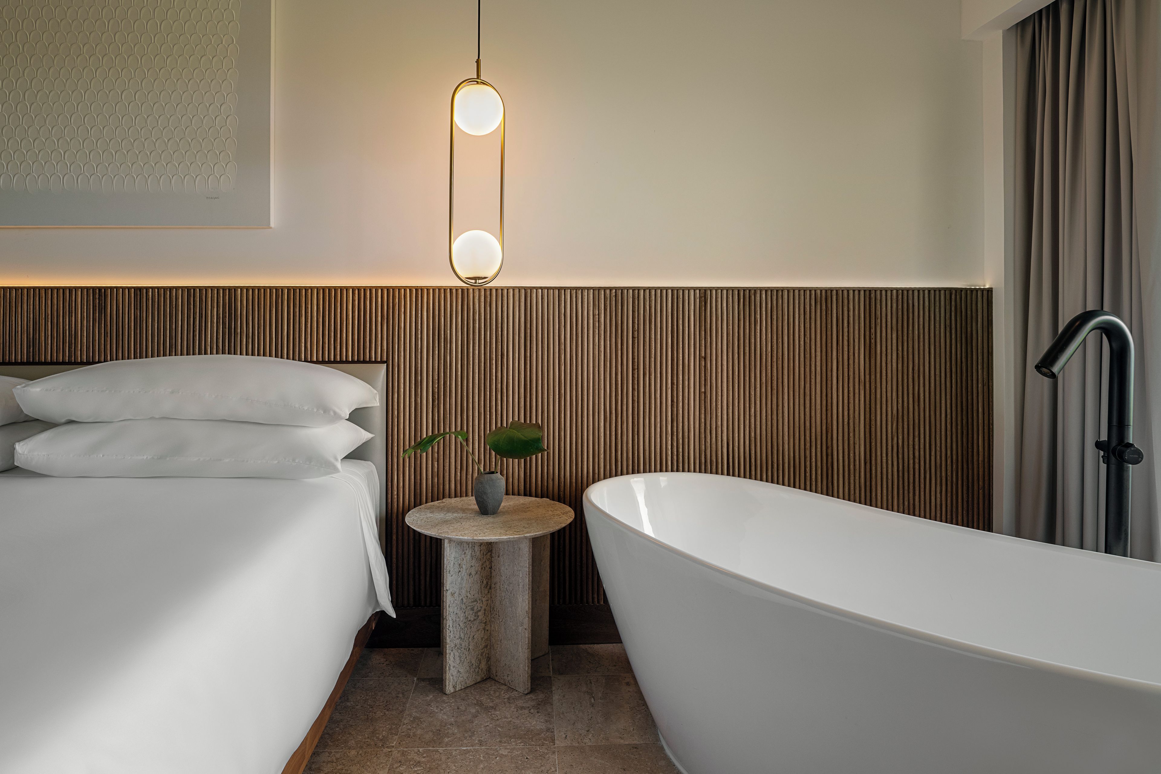 A modern bathroom with a freestanding bathtub, a bedside table with a potted plant, and a wall-mounted light fixture.