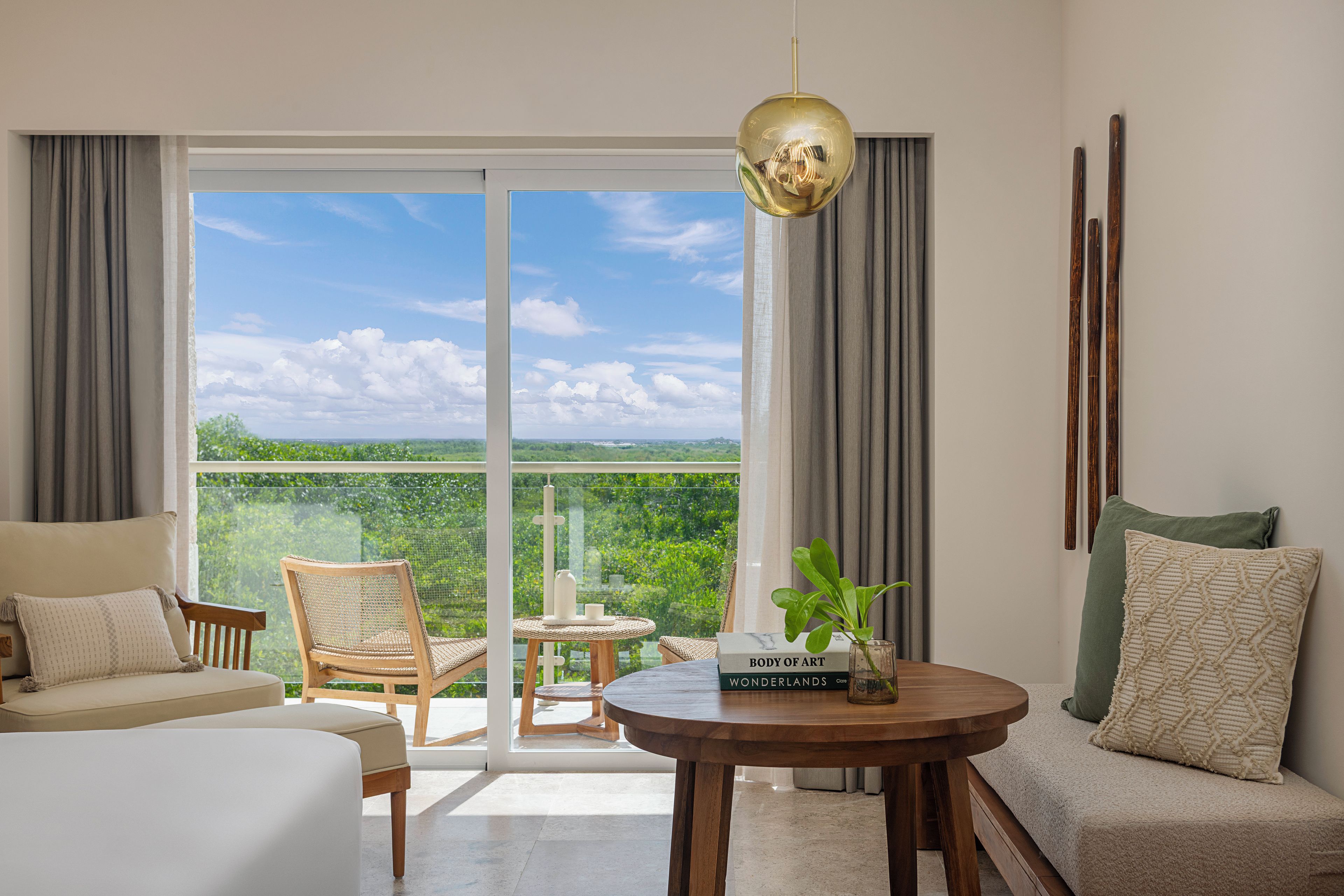 Una habitación de hotel moderna con un amplio ventanal corredero que da a un balcón, una mesa redonda de madera, un sofá y una silla.