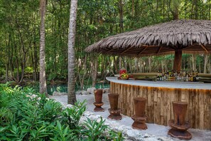 Un bar rústico al aire libre con techo de paja, rodeado de exuberante follaje tropical y una masa de agua tranquila.
