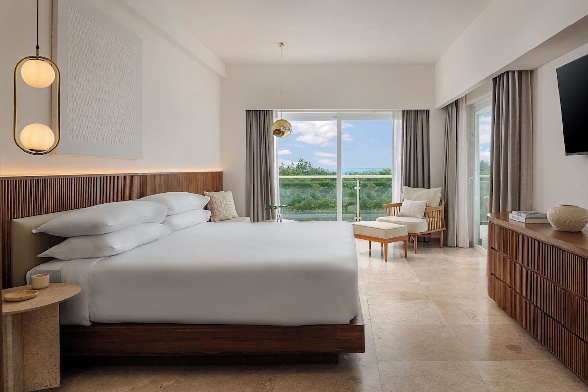 Un dormitorio moderno con una cama grande, mesitas de noche, un televisor y vistas a zonas verdes por la ventana.