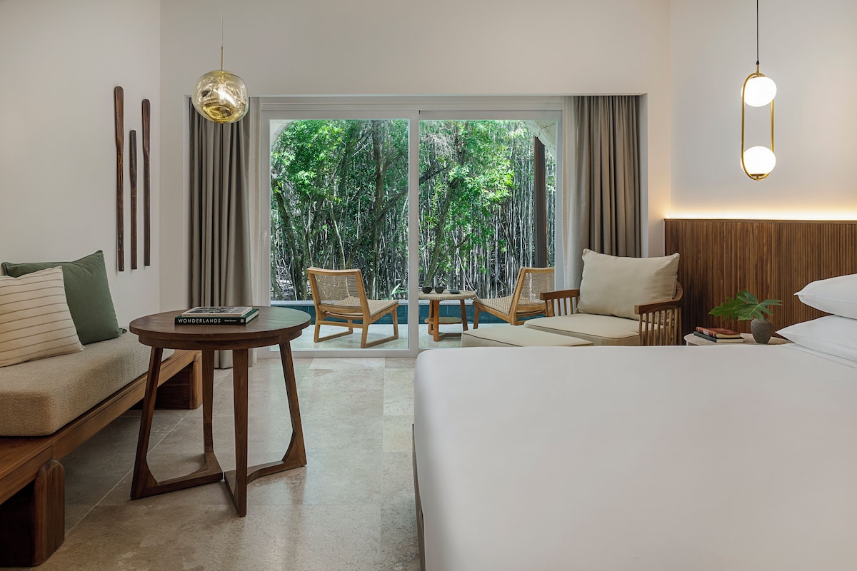 Habitación de hotel con cama, sofá, una mesita, dos sillas, lámpara colgante y vistas a la vegetación a través de la ventana.