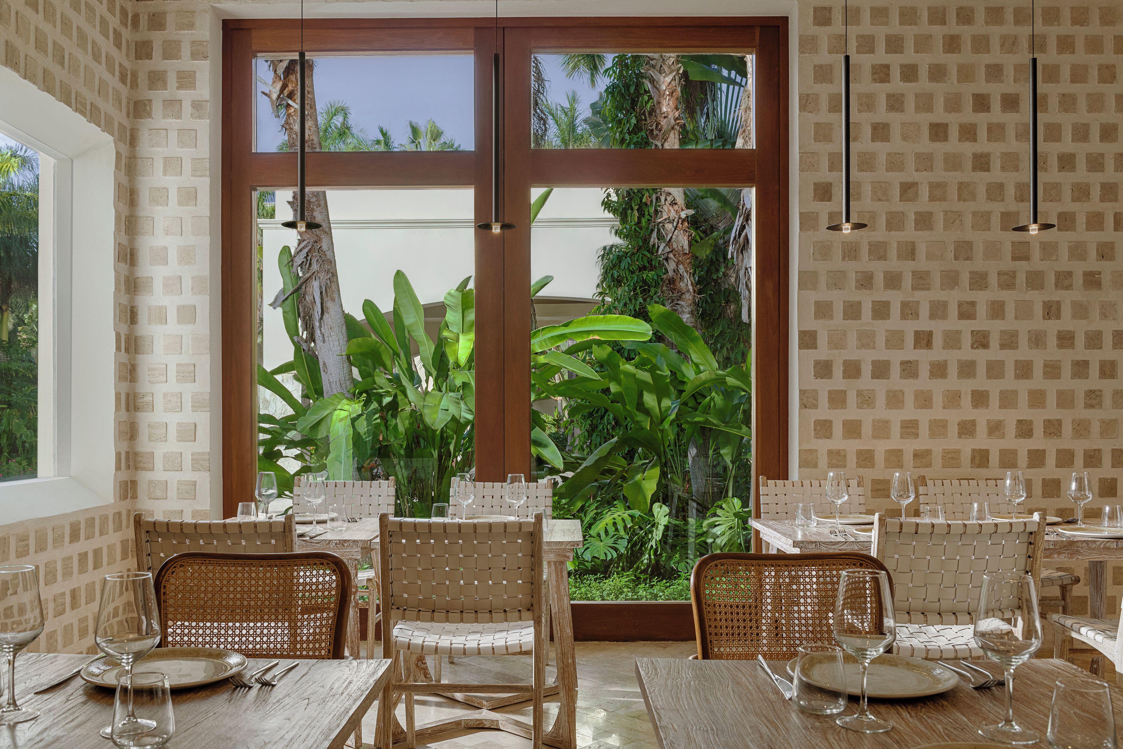 A dining area with a view of tropical plants through large windows, wicker chairs, and a brick wall pattern.