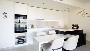 A modern kitchen with white cabinetry, a built-in oven, and a dining area with a white table and chairs. - Horizonte Benalmádena - Brand-New 3-Bedroom Apartment (Benalmádena)