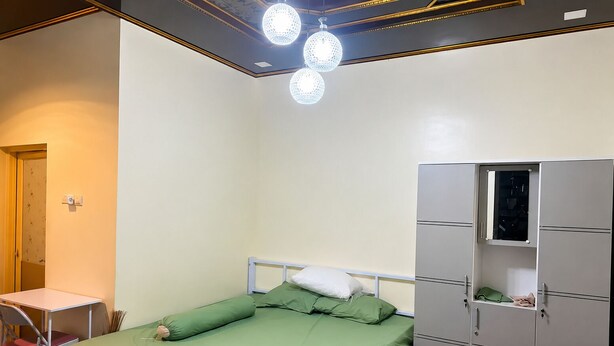 A single bed with green bedding, a white pillow, and a metal headboard. There is a small desk with a chair, a wardrobe with multiple compartments, and a ceiling light fixture. - Cozzy FK Heliconia (Jonggol)