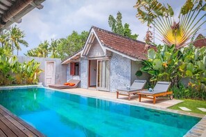 Zona de piscina con un pequeño edificio, sillas de descanso y plantas tropicales.