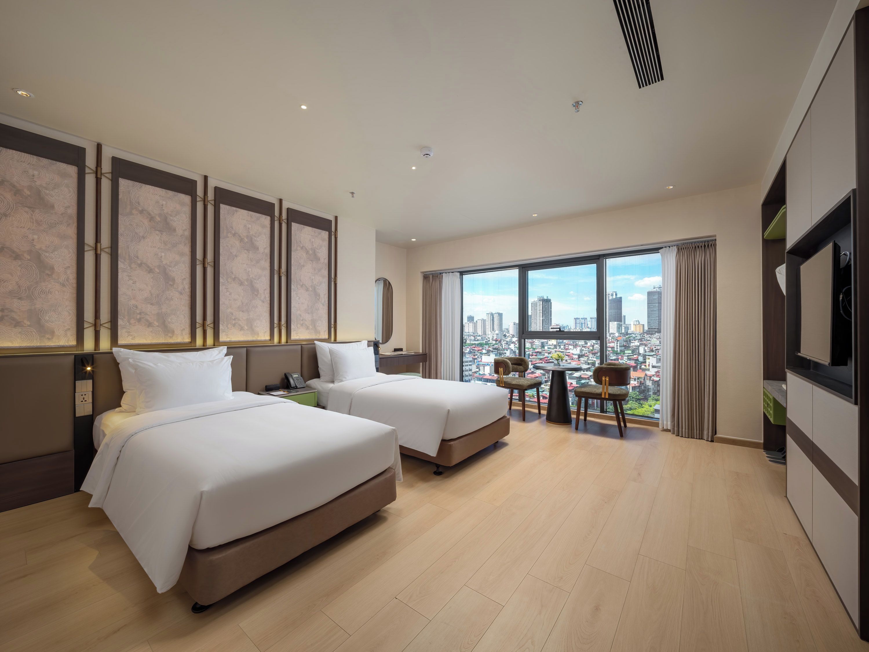 A hotel room with two beds, a city view, a dining area with chairs, and a TV on the wall.