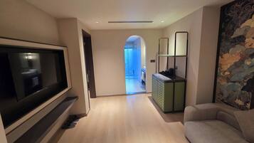 Una sala de estar moderna con un televisor de pantalla plana, un sofá y una mesa de centro. Se observa un pasillo que conduce a un baño con puerta de vidrio.