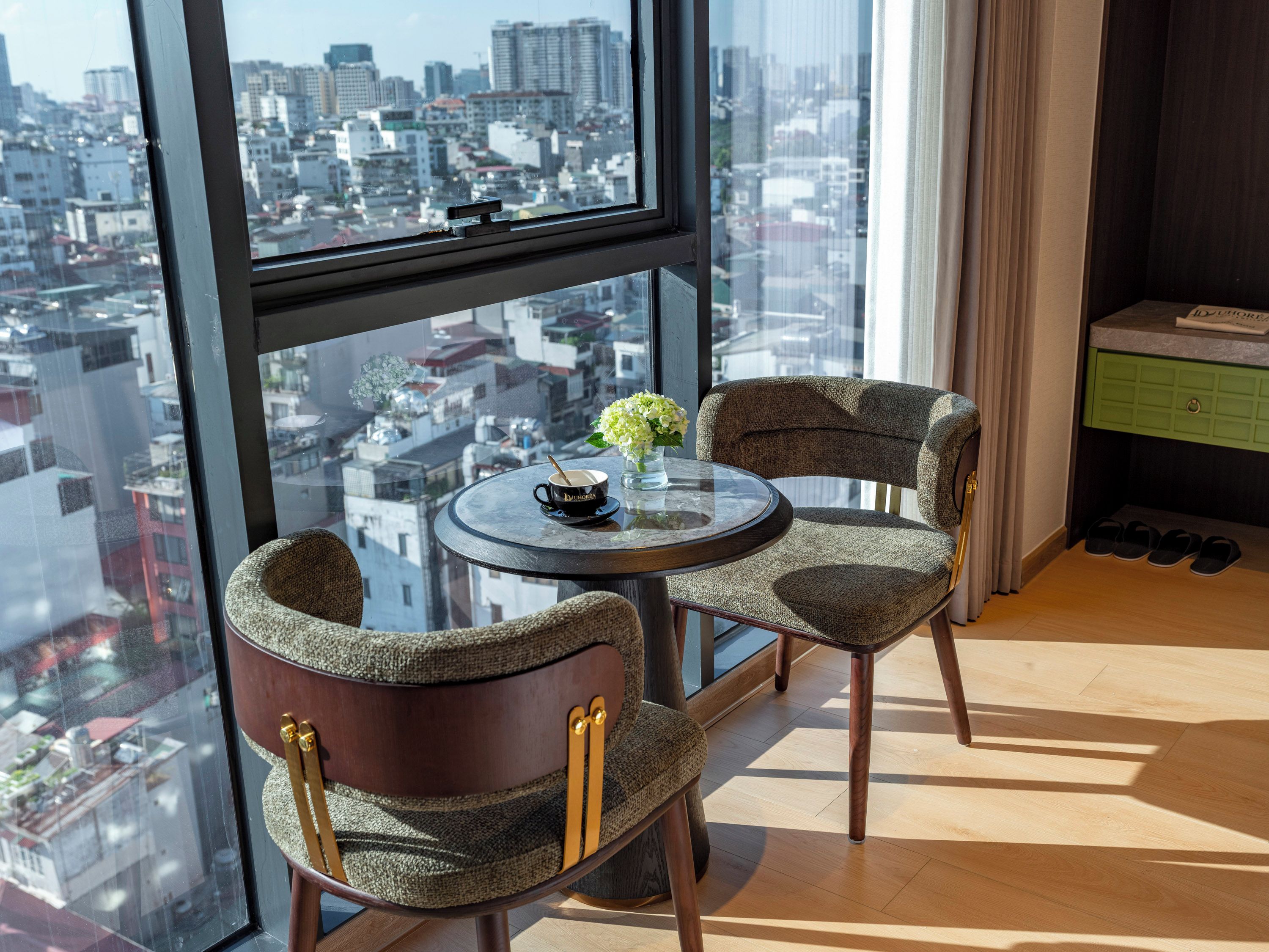 Habitación en un rascacielos con vista a la ciudad, dos sillones tapizados, una mesita redonda con una taza y flores, y un mueble verde.